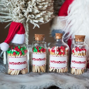 May include: Four glass bottles filled with matches, each with a cork stopper. The matches have red and green tips. Each bottle has a label that says "HOHOHO" in red letters. One bottle has a small Santa hat on top.