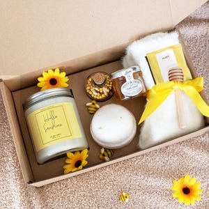 May include: A gift box with a bee theme, containing a candle, honey, bath bomb, and other items. The candle has a yellow label with the words "Hello Sunshine". The box is decorated with yellow sunflowers and bee-shaped decorations.
