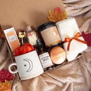 May include: A gift box filled with fall-themed items. Includes a white mug with a crescent moon and star design, a jar of matches, a candle, a bottle of room spray, a bath bomb, a pair of socks tied with an orange ribbon, and decorative fall leaves.