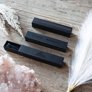 May include: A black matchbox with the text "ETHEREAL MOUNTAIN" and a mountain logo. The matchbox is open and contains black matches. The matchbox is on a wooden surface with a white feather and a pink crystal.