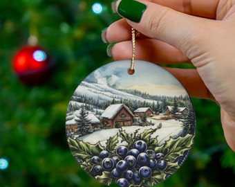 May include: A circular Christmas ornament depicting a snowy winter scene with buildings and mountains. The ornament has a gold hanging string and a cluster of blueberries and holly leaves at the bottom.