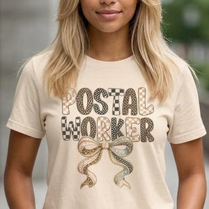 Puede incluir: Una camiseta beige con la frase "POSTAL WORKER" en una mezcla de fuentes a cuadros y estampadas. Debajo del texto hay un lazo decorativo con detalles en marrón y verde. La camiseta es de manga corta.