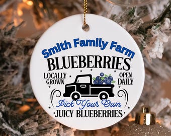 May include: A white, circular Christmas bauble with a gold hanging string. The ornament displays the text "Smith Family Farm BLUEBERRIES" in blue, with a black truck graphic carrying blueberries. The text "Locally Grown, Open Daily, Pick Your Own, Juicy Blueberries" is also included.