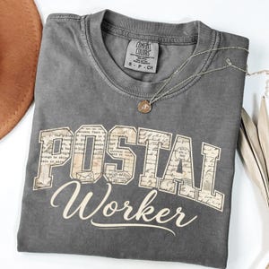 May include: A gray Comfort Colors t-shirt with the words "POSTAL WORKER" in a vintage-style font. The word "POSTAL" is in a block letter style with a collage of postage stamps. A silver necklace with a small pendant is on the shirt.