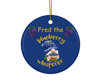 May include: A circular blue ceramic ornament with a gold hanging string. The ornament displays the text "Fred the blueberry whisperer" in yellow, and an illustration of a gnome with a basket of blueberries. Decorative blueberries are also present.