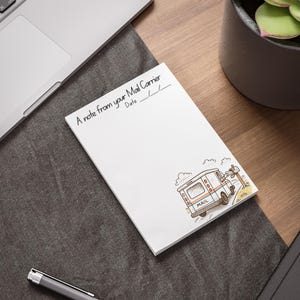 May include: White notepad with the text "A note from your Mail Carrier" and space for a date. A cartoon mail truck is in the lower right corner. The notepad is on a dark grey surface.