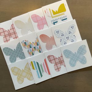 May include: A collection of white square cards with butterfly cutouts. Each butterfly has a unique patterned design in colors like pink, blue, yellow, and green. The cards are arranged on a wooden surface.