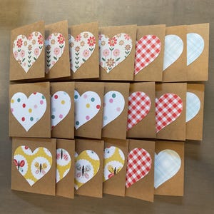 May include: A collection of handmade greeting cards with heart-shaped cutouts. The cards feature various patterns, including floral, polka dots, gingham, and butterfly designs. The cards are made of brown cardstock.