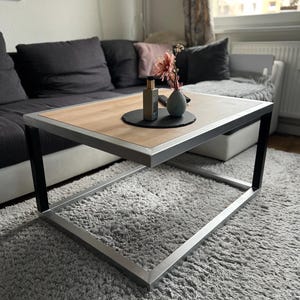 Peut inclure: Une table basse moderne avec un plateau en bois clair et un cadre en métal. La table est rectangulaire et repose sur un tapis shaggy gris. Un petit vase avec des fleurs roses et un flacon de parfum sont sur la table.