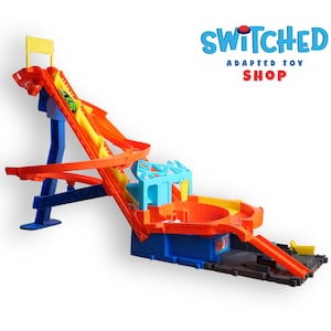 May include: A colorful toy race track set with a blue base, orange and yellow ramps, and a small green race car. The set includes the text "SWITCHED ADAPTED TOY SHOP" in blue and red.