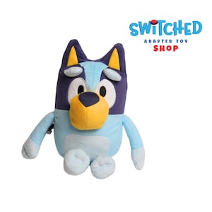 May include: A plush toy dog in light blue, dark blue, and yellow. The dog has large, round eyes and a yellow nose. The toy is sitting with its arms and legs outstretched. The text "SWITCHED ADAPTED TOY SHOP" is in the upper right corner.