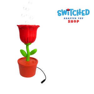 May include: A red plastic flower-shaped bubble machine with a green stem and leaves, set in an orange pot. Bubbles float above the flower. The words "Switched Adapted Toy Shop" are in the upper right corner.