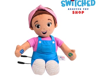 Peut inclure: Une poupée en peluche avec des cheveux bruns, un bandeau rose et un t-shirt rose sous une salopette bleue. La poupée a des notes de musique et du texte sur les bras et les pieds. Le logo Switched Adapted Toy Shop est en haut à droite.