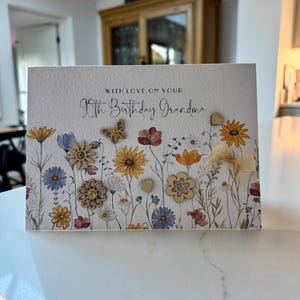May include: A white card with a floral design and the text "With love on your 90th Birthday Grandma". The card has small wooden embellishments in the shape of flowers, hearts, and butterflies.