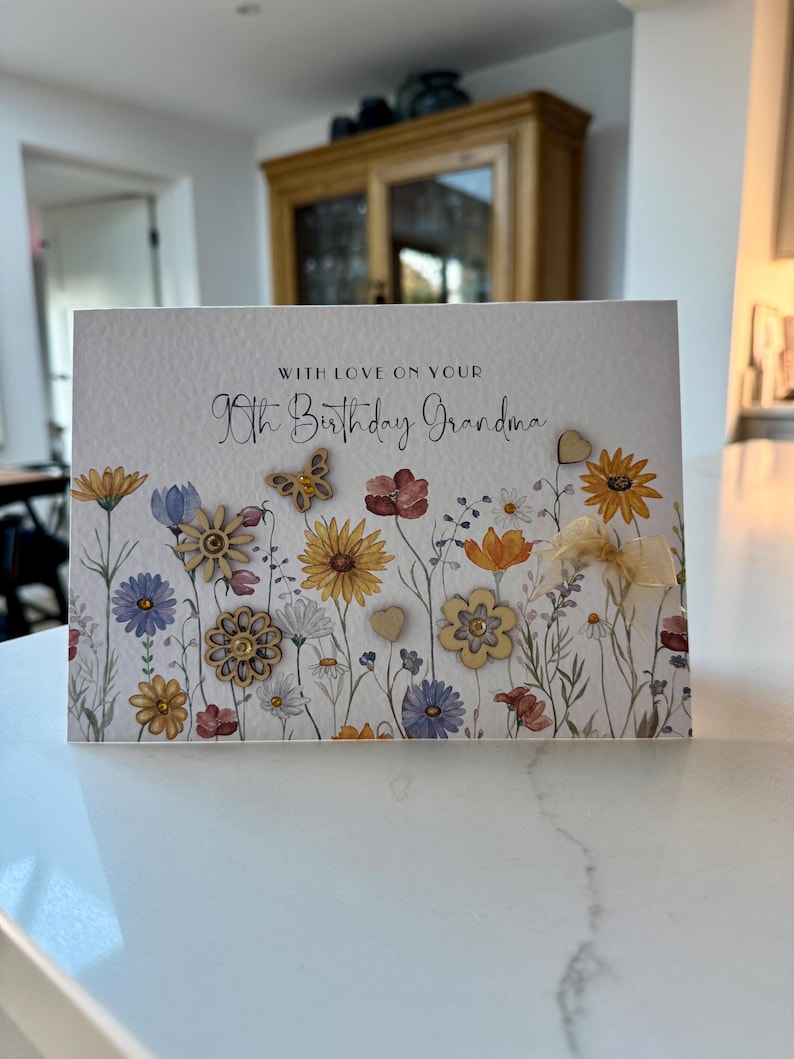 May include: A white card with a floral design and the text "With love on your 90th Birthday Grandma". The card has small wooden embellishments in the shape of flowers, hearts, and butterflies.