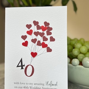 Puede incluir: Tarjeta de aniversario blanca con un diseño de globos en forma de corazón rojos y granates. El número "40" es granate, con el texto "with love to my amazing Husband on our 40th Wedding Anniversary".