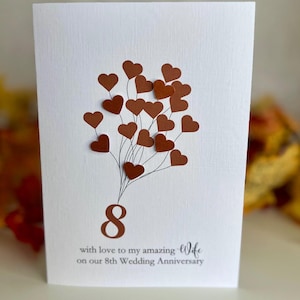 Può includere: Biglietto bianco con ritagli a forma di cuore marrone disposti come palloncini. Il numero 8 e la scritta «con amore alla mia fantastica moglie per il nostro 8° anniversario di matrimonio» sono stampati in marrone. Biglietto di anniversario.