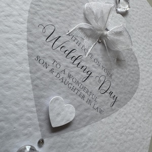 May include: A white wedding card with a heart-shaped design. The card features the words "Wedding Day" in elegant script, along with a white bow and decorative crystals. The card reads "With love on your Wedding Day to a wonderful Son & Daughter in Law."