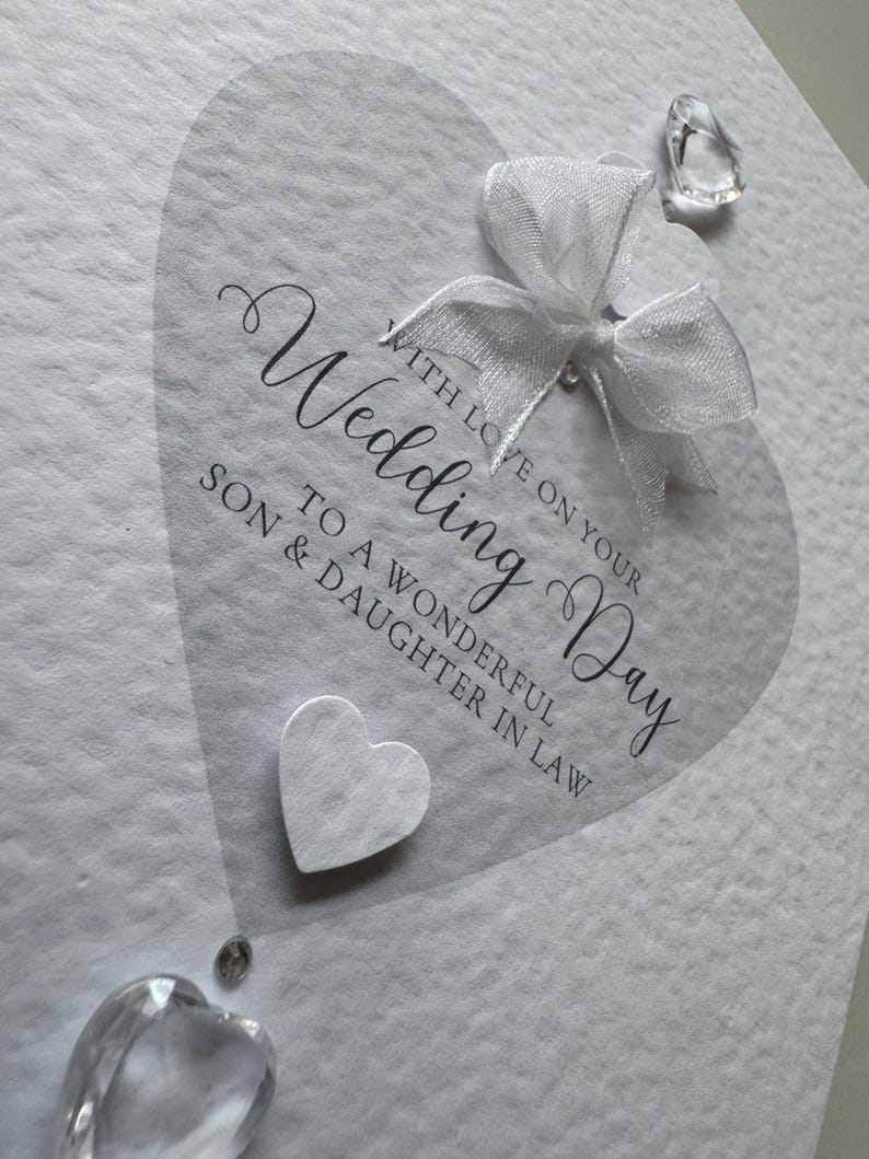May include: A white wedding card with a heart-shaped design. The card features the words "Wedding Day" in elegant script, along with a white bow and decorative crystals. The card reads "With love on your Wedding Day to a wonderful Son & Daughter in Law."
