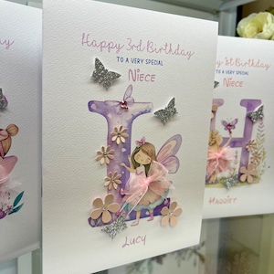 May include: A handmade birthday card with a purple watercolor letter "L" decorated with pink flowers, glitter butterflies, and a fairy illustration. The card says "Happy 3rd Birthday to a very special Niece" and is signed "Lucy".
