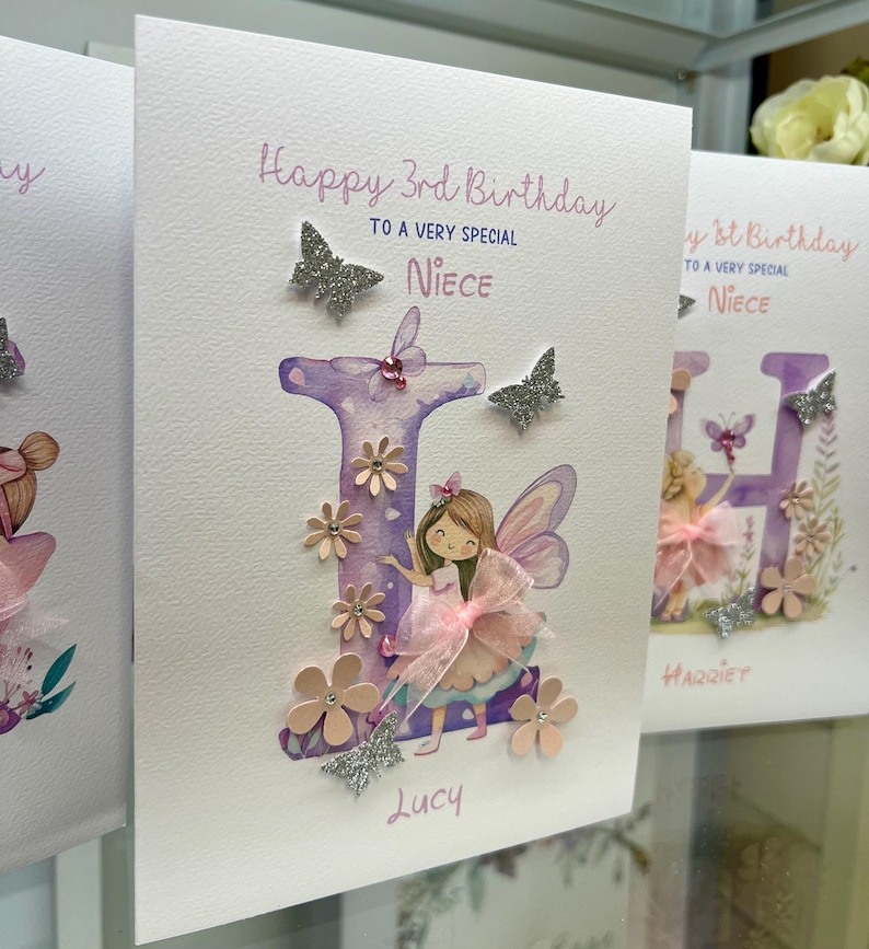 May include: A handmade birthday card with a purple watercolor letter "L" decorated with pink flowers, glitter butterflies, and a fairy illustration. The card says "Happy 3rd Birthday to a very special Niece" and is signed "Lucy".