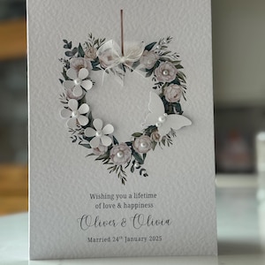 May include: A wedding card with a heart-shaped floral wreath. The wreath features white flowers, pearls, and foliage. A white butterfly is on the right. The card reads: "Wishing you a lifetime of love & happiness, Oliver & Olivia, Married 24th January 2025."