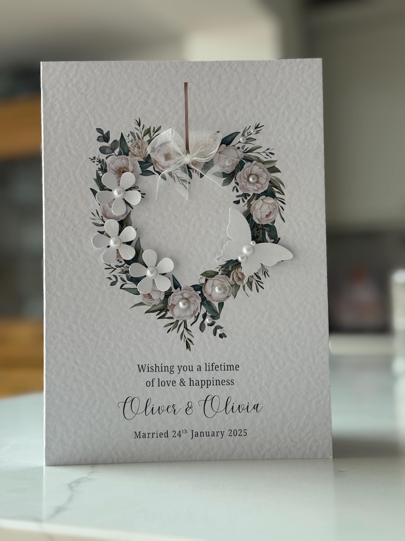 May include: A wedding card with a heart-shaped floral wreath. The wreath features white flowers, pearls, and foliage. A white butterfly is on the right. The card reads: "Wishing you a lifetime of love & happiness, Oliver & Olivia, Married 24th January 2025."