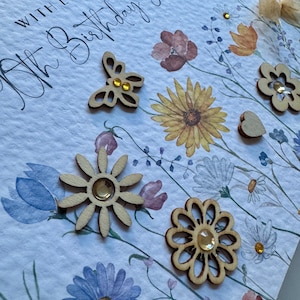 May include: A white card with the text "With love on your 80th Birthday Grandma" in black cursive writing. The card is decorated with watercolor flowers and wooden embellishments, including a butterfly, a flower, and a heart.