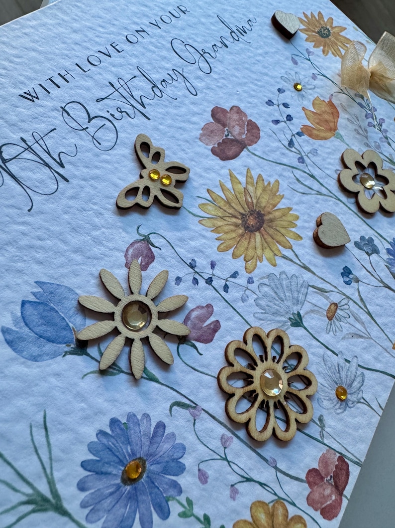 May include: A white card with the text "With love on your 80th Birthday Grandma" in black cursive writing. The card is decorated with watercolor flowers and wooden embellishments, including a butterfly, a flower, and a heart.
