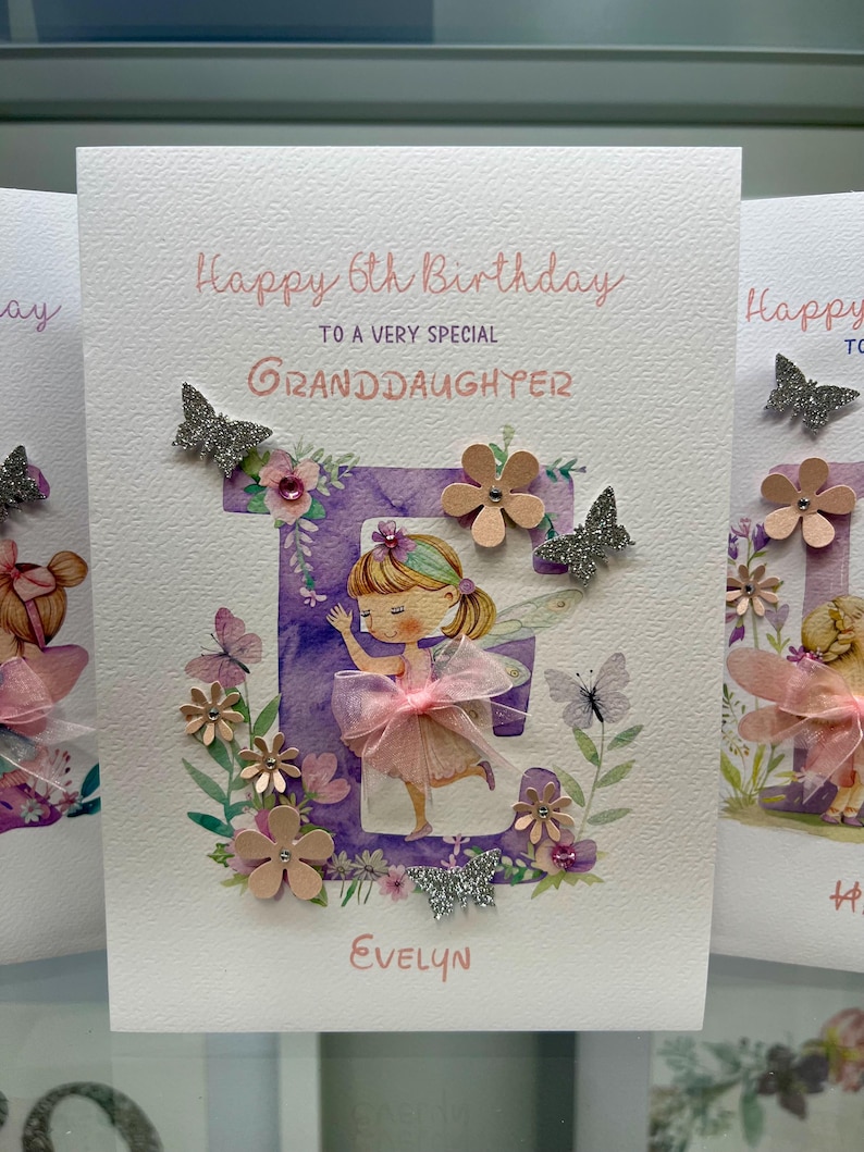 May include: A purple and white card with a pink and white fairy illustration and the text "Happy 6th Birthday to a very special Granddaughter Evelyn". The card is decorated with glitter butterflies, flowers, and rhinestones.