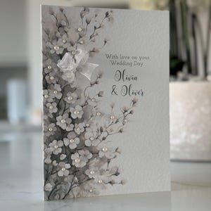 Może przedstawiać: Biała kartka z akwarelowym wzorem kwiatowym i perłowymi ozdobami. Na kartce widnieje napis "With love on your Wedding Day Olivia & Oliver".