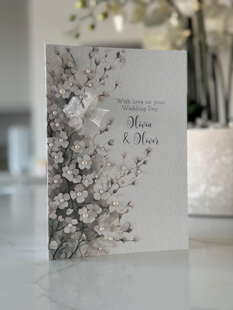Może przedstawiać: Biała kartka z akwarelowym wzorem kwiatowym i perłowymi ozdobami. Na kartce widnieje napis "With love on your Wedding Day Olivia & Oliver".