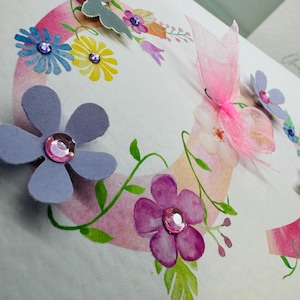 May include: A handmade card with a pink and white watercolour background featuring a floral wreath with butterflies and rhinestones. The card is perfect for a birthday or other special occasion.