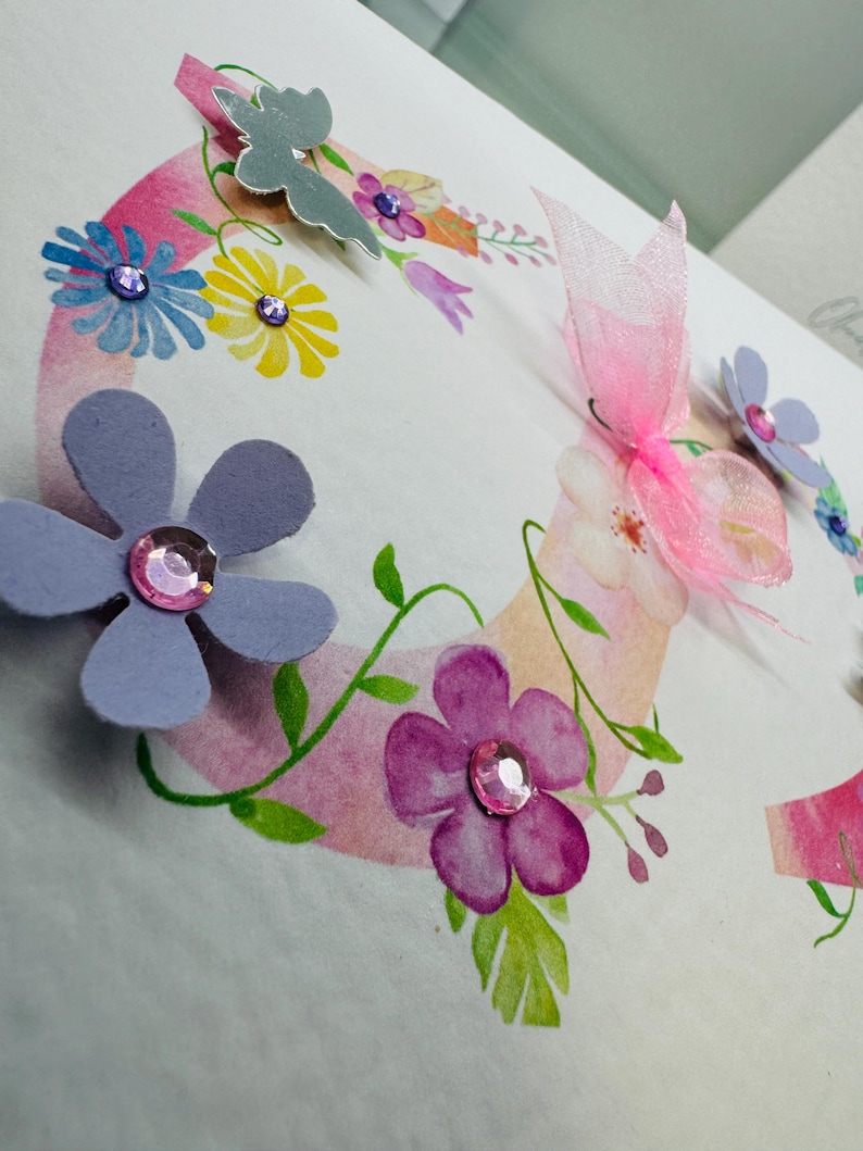 May include: A handmade card with a pink and white watercolour background featuring a floral wreath with butterflies and rhinestones. The card is perfect for a birthday or other special occasion.