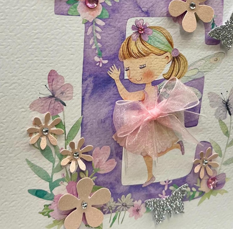 May include: A purple letter E with a watercolor design and a fairy with pink wings and a pink bow. The letter is surrounded by flowers and butterflies.