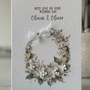 May include: A white wedding card with a floral wreath design. The wreath features white flowers, pearls, wooden hearts, and green leaves. The card reads "With Love on Your Wedding Day" and the names "Olivia & Oliver".