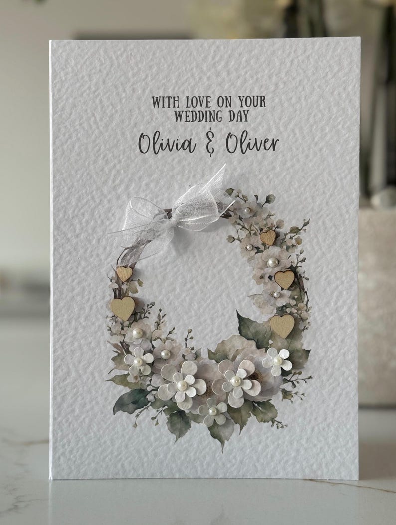 May include: A white wedding card with a floral wreath design. The wreath features white flowers, pearls, wooden hearts, and green leaves. The card reads "With Love on Your Wedding Day" and the names "Olivia & Oliver".