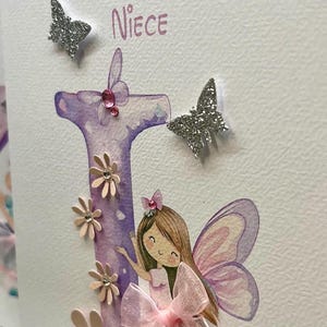 May include: A handmade card with a purple letter "I" decorated with pink flowers and a fairy with pink wings. The card says "To Niece" at the top.