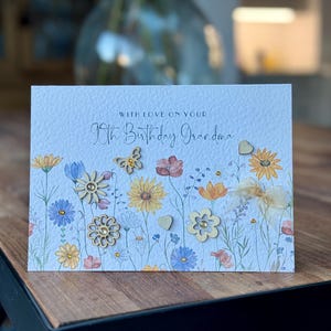 May include: A light blue card with a floral design and the text "With love on your 90th Birthday Grandma". The card has wooden embellishments in the shape of flowers and a butterfly.