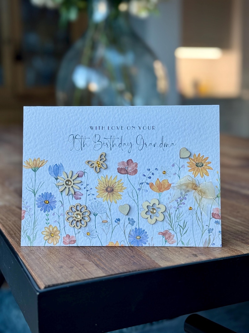 May include: A light blue card with a floral design and the text "With love on your 90th Birthday Grandma". The card has wooden embellishments in the shape of flowers and a butterfly.