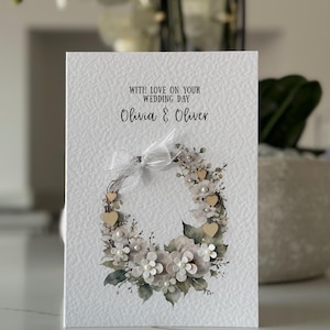 May include: A wedding card with the text "WITH LOVE ON YOUR WEDDING DAY Olivia & Oliver." The card features a floral wreath design with white flowers, wooden hearts, and pearls. A white ribbon is tied at the top.