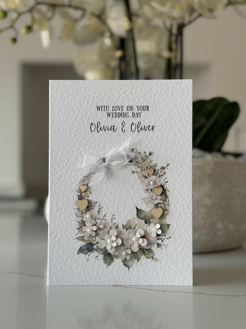 May include: A wedding card with the text "WITH LOVE ON YOUR WEDDING DAY Olivia & Oliver." The card features a floral wreath design with white flowers, wooden hearts, and pearls. A white ribbon is tied at the top.