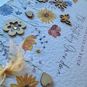 May include: A white card with a floral design and the text "Happy Birthday" and "With love on your Birthday". The card is decorated with wooden hearts, flowers, and a butterfly.