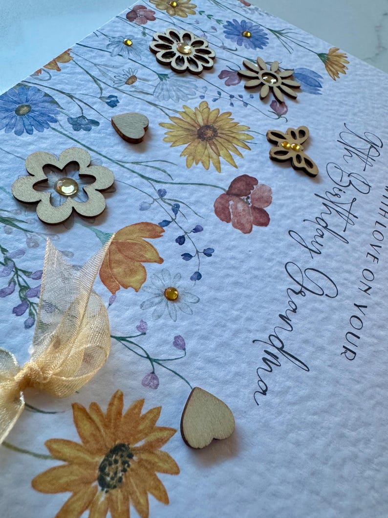 May include: A white card with a floral design and the text "Happy Birthday" and "With love on your Birthday". The card is decorated with wooden hearts, flowers, and a butterfly.