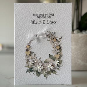 May include: A wedding card with the text "WITH LOVE ON YOUR WEDDING DAY" and the names "Olivia & Oliver". The card features a floral wreath design with white and gray flowers, wooden hearts, and pearls. A white ribbon is tied at the top.