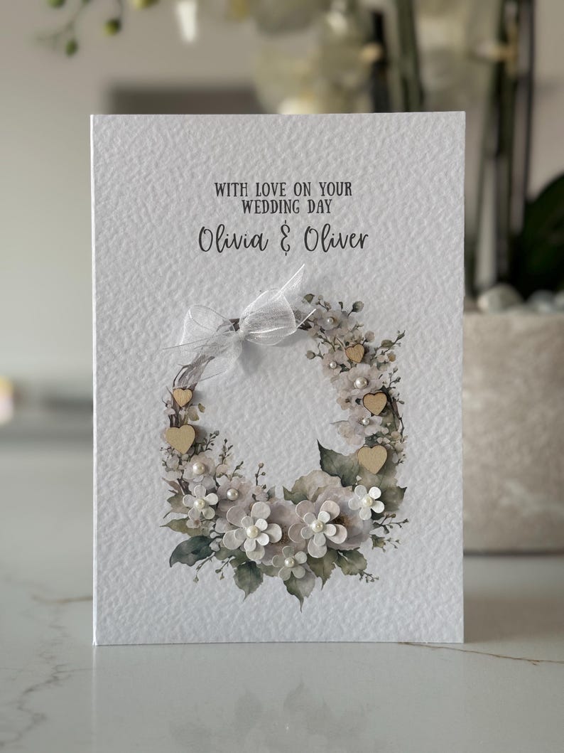 May include: A wedding card with the text "WITH LOVE ON YOUR WEDDING DAY" and the names "Olivia & Oliver". The card features a floral wreath design with white and gray flowers, wooden hearts, and pearls. A white ribbon is tied at the top.