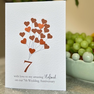 May include: White anniversary card featuring a heart-shaped balloon design in brown and orange. The number 7 is displayed, with the text "with love to my amazing Husband on our 7th Wedding Anniversary."