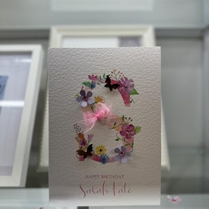 May include: A white card with a pink and purple floral design in the shape of the letter "S". The card says "Happy Birthday Sarah Kate" in pink lettering.