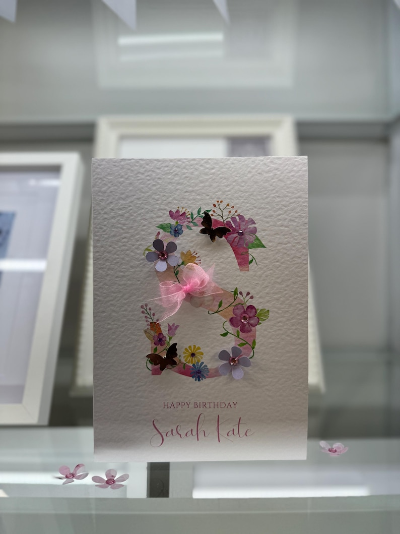 May include: A white card with a pink and purple floral design in the shape of the letter "S". The card says "Happy Birthday Sarah Kate" in pink lettering.