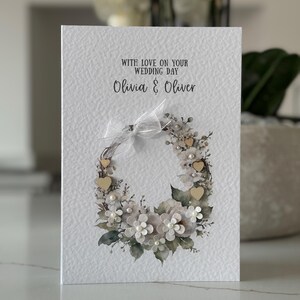 May include: A wedding card with a textured white background. The card features a floral wreath design with white flowers, pearls, and small wooden hearts. The text "WITH LOVE ON YOUR WEDDING DAY" and the names "Olivia & Oliver" are printed on the card.